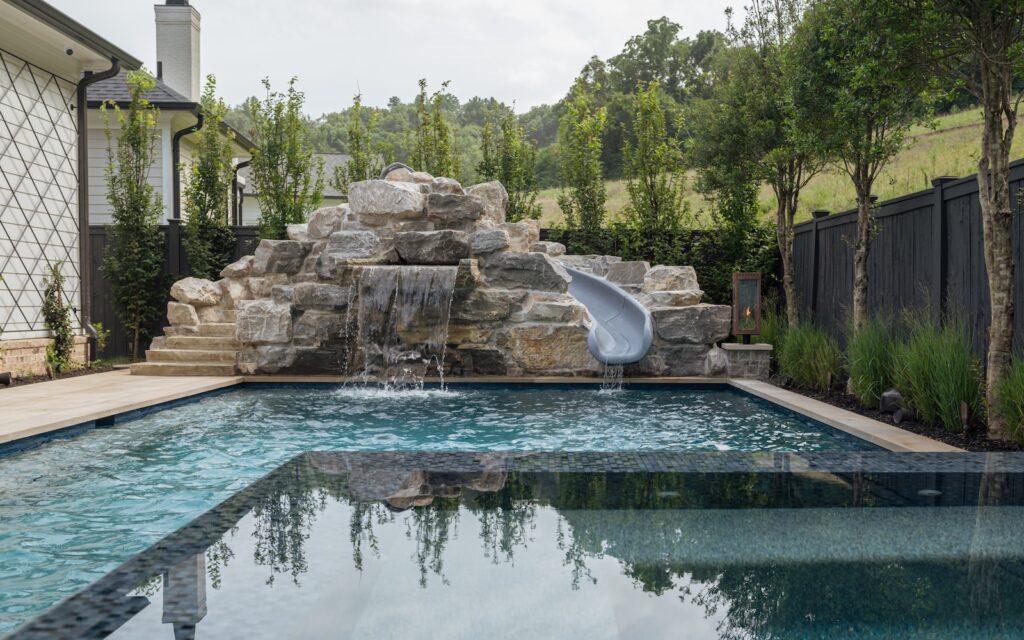 Tranquil pool & spa with sculptural water feature, water slide, tree-lined fencing | Milosi Nashville Landscaping